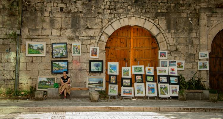 Art market display on historic stone wall with framed paintings and a seated vendor.