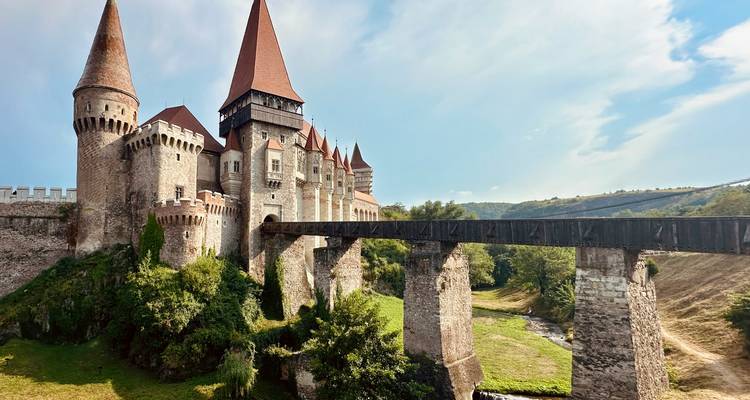 Impressive medieval Corvin Castle with tall red-roofed towers and long wooden bridge over green valley.