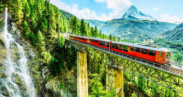Rode bergtrain steekt hoge brug over met waterval en Matterhorn die in de verte uittorent.