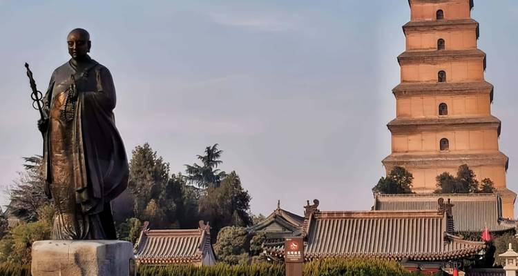 Una estatua de bronce de un monje budista se encuentra frente a la histórica Pagoda del Gran Ganso Salvaje de varios pisos, enmarcada por los techos tradicionales del templo y árboles bajo un cielo despejado.