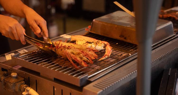 Gros plan sur des mains faisant griller de grandes crevettes sur une grille à charbon de bois métallique dans un étal de restauration.