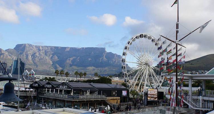 Cape Town’s V&A Waterfront with ferris wheel, shops, flag masts and Table Mountain backdrop.