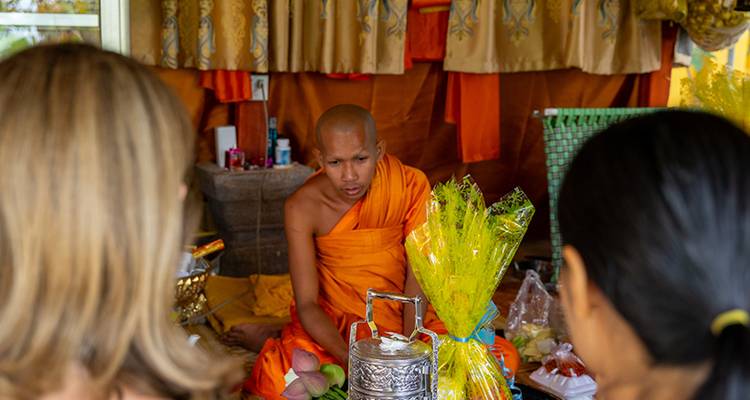 Buddhist monk in orange robe performing a blessing ceremony inside a small shrine with visitors watching.