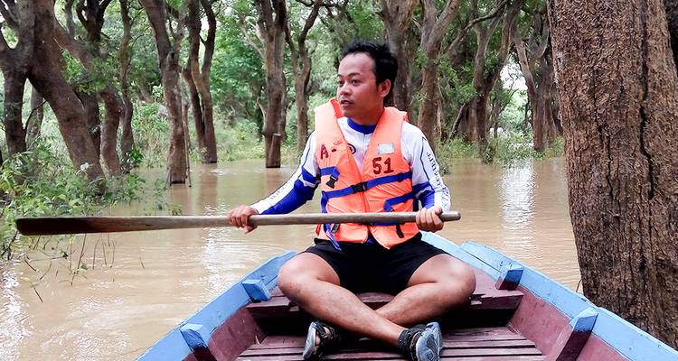 Man wearing life vest paddling a small wooden boat through flooded forest waters.