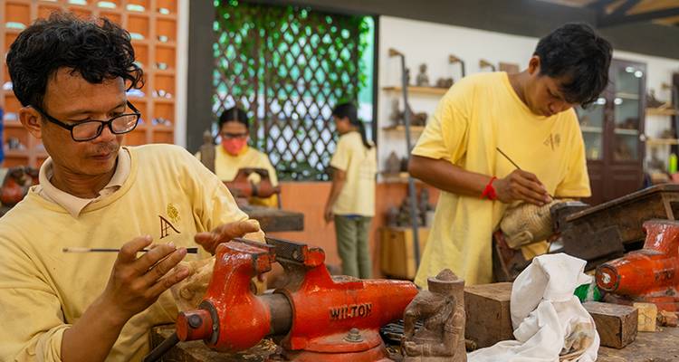 Artisans in a workshop carefully carving and sanding pieces at cluttered wooden benches.