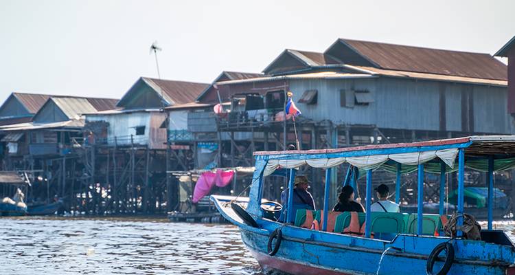 Blue long-tail boat carrying passengers past stilt houses over a wide lake.