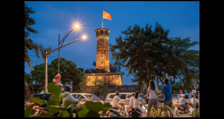 Scène du soir de la Tour du Drapeau de Hanoï illuminée par le drapeau vietnamien tandis que la circulation routière s'estompe en dessous.