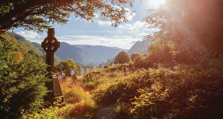 Ancient Celtic cross in sunlit Glendalough cemetery framed by lush valley and distant mountains.