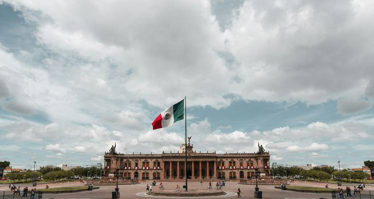 Vaste place avec le drapeau mexicain flottant au-dessus d'un bâtiment gouvernemental de style néoclassique sous des nuages dramatiques.