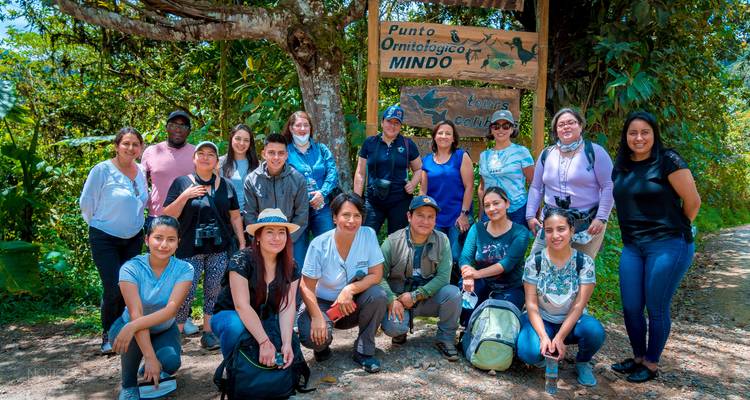 Large tour group posing by a wooden sign reading ‘Mindo’ in lush forest surroundings.