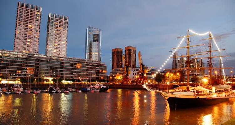 Vista nocturna de la costa de Puerto Madero con un velero histórico iluminado y el moderno perfil de Buenos Aires reflejándose en el río.