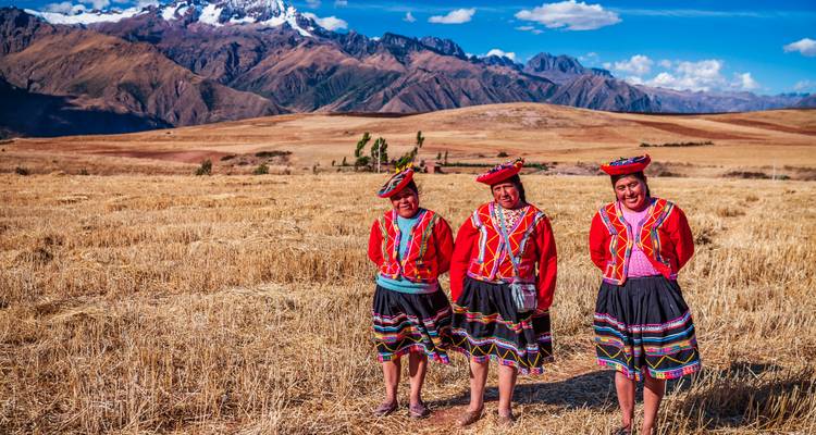 Three women in vivid traditional Andean dress standing in a golden field with snow-capped mountains behind