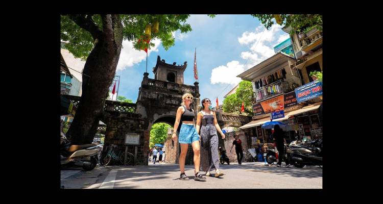 Zwei weibliche Reisende gehen an einem alten Stadttor auf einer lebhaften Straße in Hanoi unter einem strahlend blauen Himmel vorbei