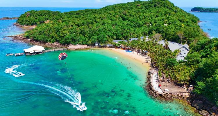 Aerial view of a turquoise bay and sandy beach where a speedboat tows a parasail against lush green island