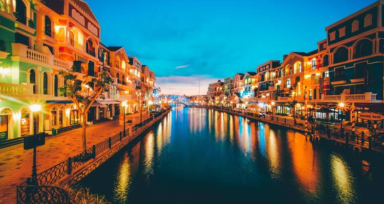 Evening view of a lit canal flanked by colorful facades reflecting in calm water against a deep blue sky