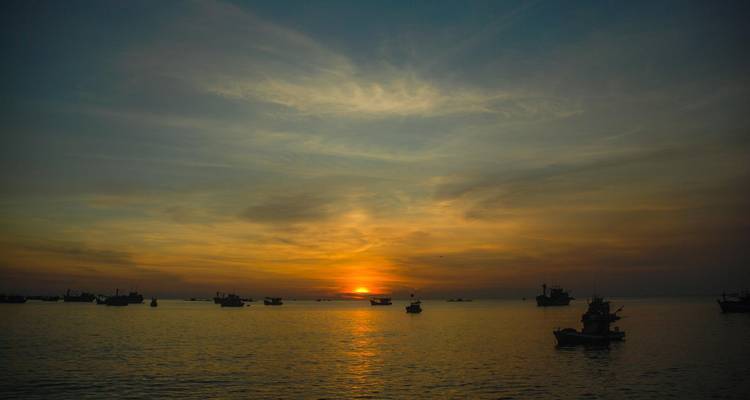 Serene sunset over a calm sea dotted with small fishing boats and a glowing orange horizon