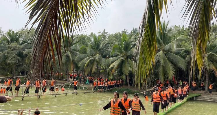 Eine große Gruppe von Jugendlichen in orangefarbenen Rettungswesten balanciert über Seilbrücken, die sich über einen grünen Teich spannen.