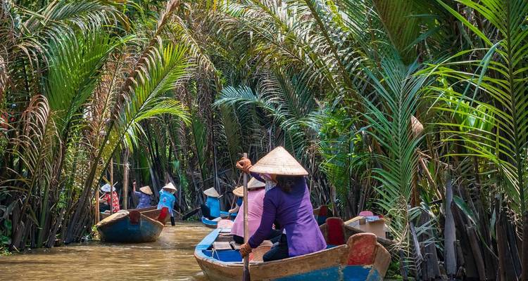 Ruderer in kegelförmigen Hüten paddeln blaue Holzboote durch enge Wasserwege mit Nipa-Palmen.
