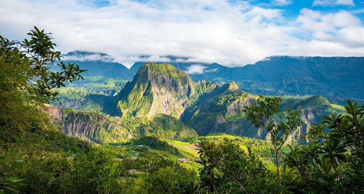 Verdant volcanic peak rises under a patchy sky, framed by tropical foliage.