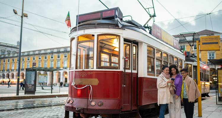 Gruppe von Freunden, die neben einer traditionellen roten Straßenbahn auf dem Hauptplatz von Lissabon an einem bewölkten Nachmittag auf einem Telefon schauen