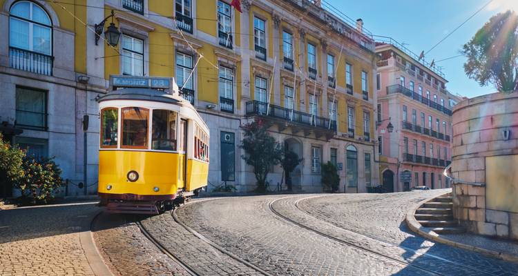 Ikonische gelbe Straßenbahn, die eine gekrümmte Kopfsteinpflasterstraße entlang an pastellfarbenen Gebäuden im alten Viertel Lissabons hinaufführt
