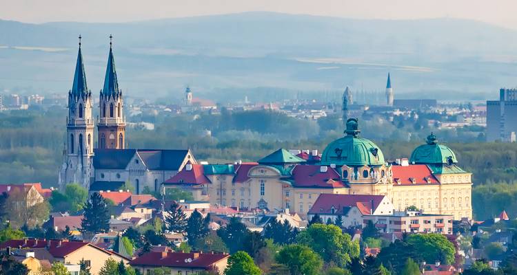 Panoramablick auf die Stadt mit Doppelkirchentürmen und Kuppeldächern des Palastes, eingebettet in grüne Hügellandschaften.
