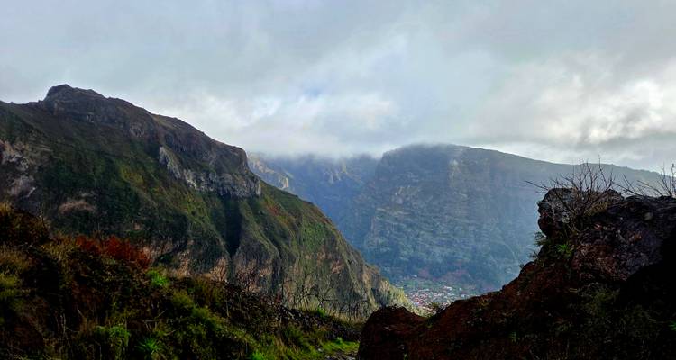 Des crêtes couronnées de nuages avec des traces d'un village en contrebas, vues au-delà du premier plan rocheux.