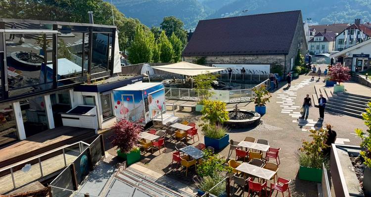 Cour ensoleillée avec tables et chaises, petits commerces alimentaires et petits bassins dans un cadre en pente.