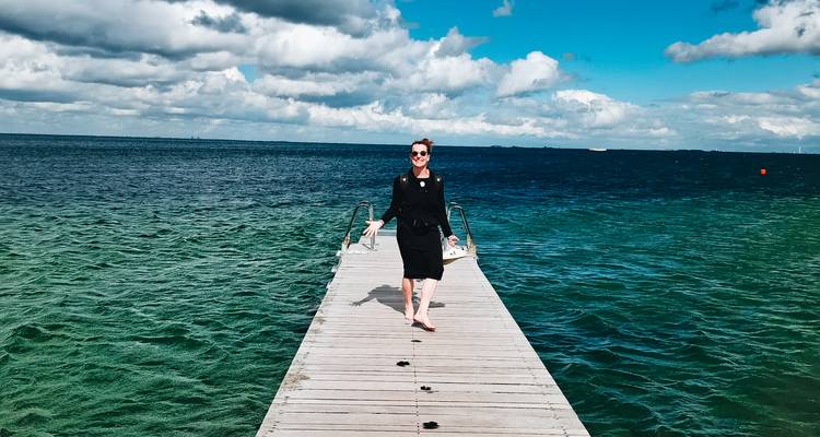 Femme souriante marchant pieds nus sur une jetée en bois s'étendant dans une mer bleu-vert cristalline