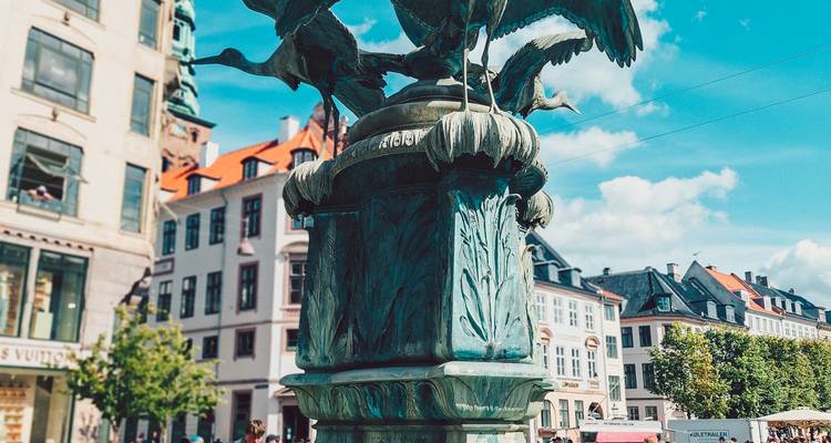 Fuente de cigüeña de bronce en una plaza de la ciudad con edificios históricos coloridos y un cielo azul brillante de fondo.