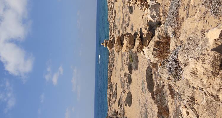Stacked stone cairn on a dry coastal plain with blue sea and boat wake beyond.