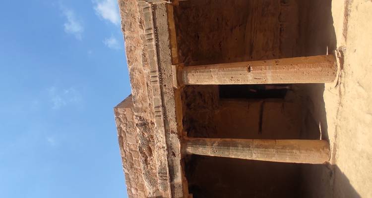 Sandstone façade with two columns of an excavated tomb courtyard under a bright blue sky.