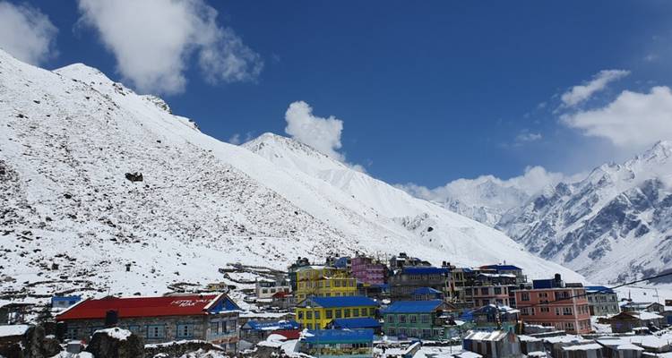 Colorful village buildings nestled in a snowy Himalayan valley beneath towering white-capped peaks and a deep blue sky.