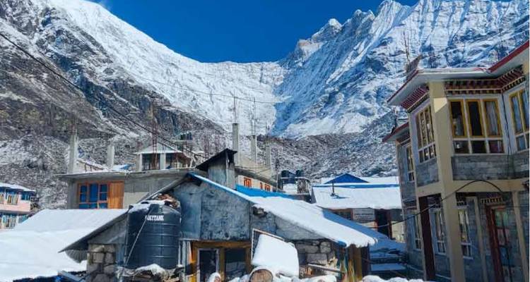 Stone and timber houses of a small mountain settlement sit under steep, snow-covered cliffs and a clear sky.