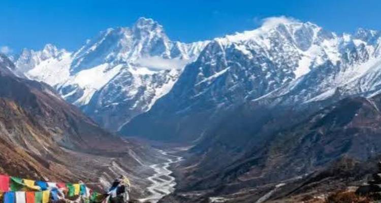 Prayer flags flutter above a dramatic V-shaped valley lined by jagged snowy peaks with a braided river below.