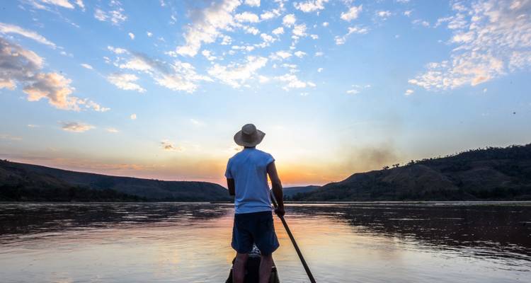 Traveller in a canoe paddles toward a fiery sunset on a wide tranquil river flanked by hills.