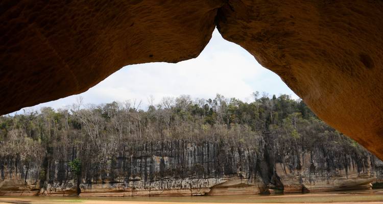 View from a cave-like rock arch framing a high cliff wall and forest across a river.