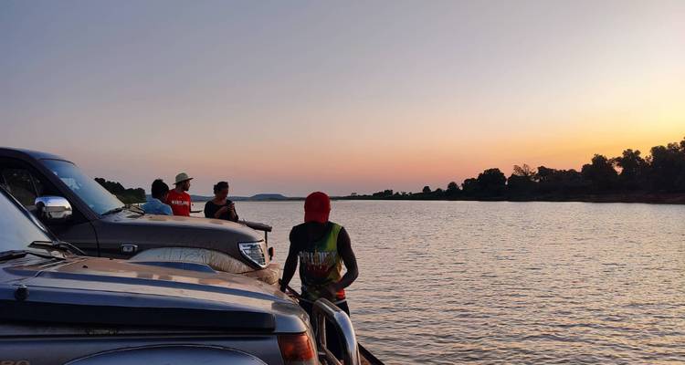 Travellers stand beside 4x4 vehicles on a river ferry at dusk, watching the pastel sunset.
