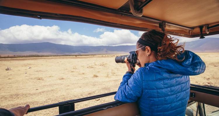 Een vrouw in een safariauto die uit het raam leunt met een telefotocamera om wilde dieren op een open vlakte te fotograferen.