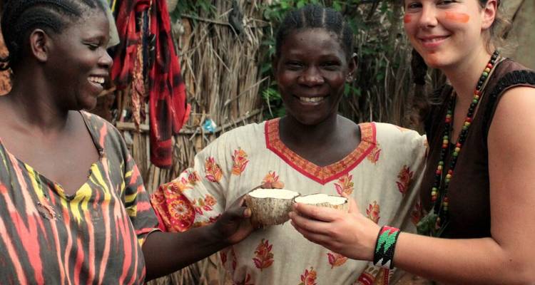 Drie vrouwen - twee locals en één reizigster - die lachend met elkaar aanstoten met calabash-bekers voor een rieten hut.