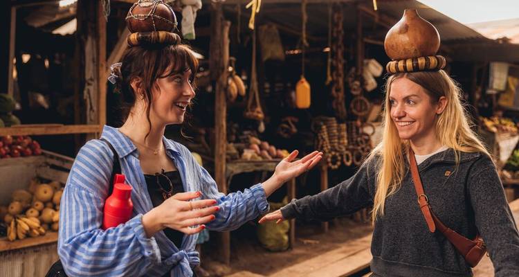 Twee vrouwelijke reizigers die houten kalebassen op hun hoofd balanceren en lachen op een openluchtgroenten markt.