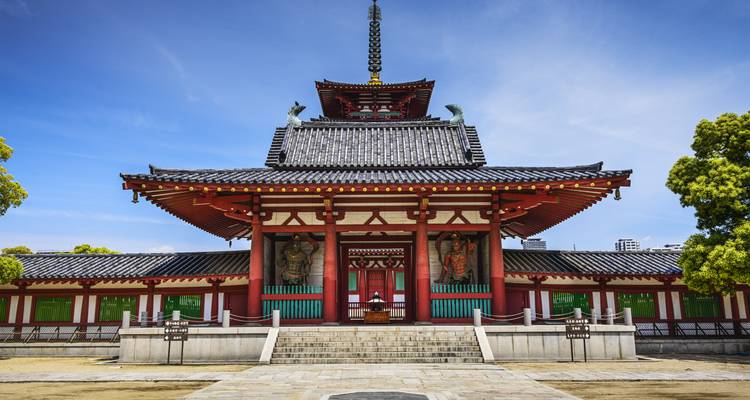 Vista simétrica del salón del templo Shitenno-ji con pilares rojos y estatuas guardianas bajo un cielo azul.
