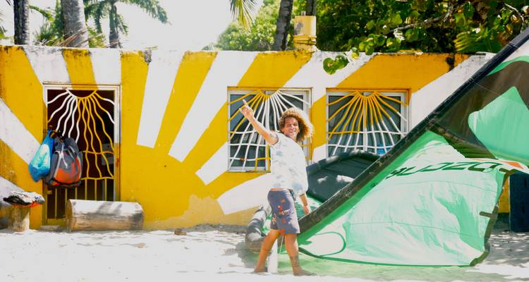 Kitesurfeur souriant levant la main près d'une cabine de plage jaune vif et d'une voile verte