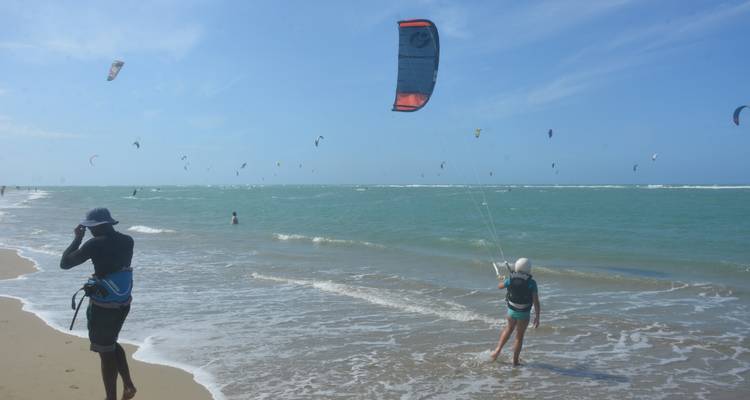 Kitesurfeur en apprentissage avec instructeur en eau peu profonde, de nombreux cerfs-volants colorés parsemant le ciel le long d'un littoral vaste.