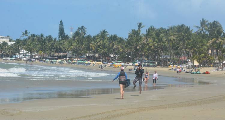 Plage bordée de cocotiers avec des vacanciers, des cerfs-volants dans le ciel et du sable mouillé reflétant la ville remplie de palmiers.