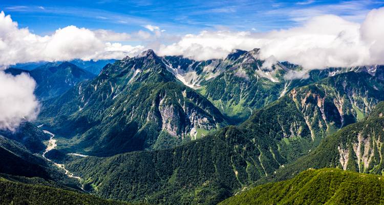 Panoramablick auf die japanischen Alpen mit tiefgrünen Tälern und Wolken, die sich an die Gipfel schmiegen.