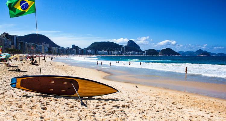 Día soleado en la Playa de Copacabana con tabla de surf, bandera brasileña, bañistas, horizonte de la ciudad y montañas.