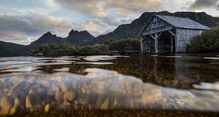 Historic wooden boat shed on Dove Lake with Cradle Mountain peaks under dramatic clouds, half-submerged perspective.