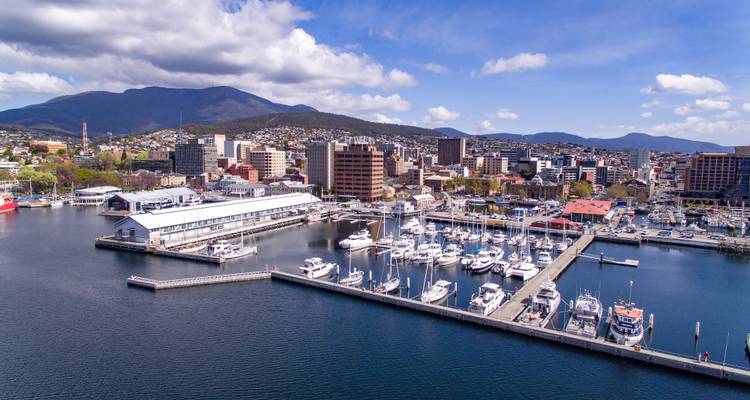 Aerial harbour scene with marinas, city buildings and mountain backdrop under scattered clouds