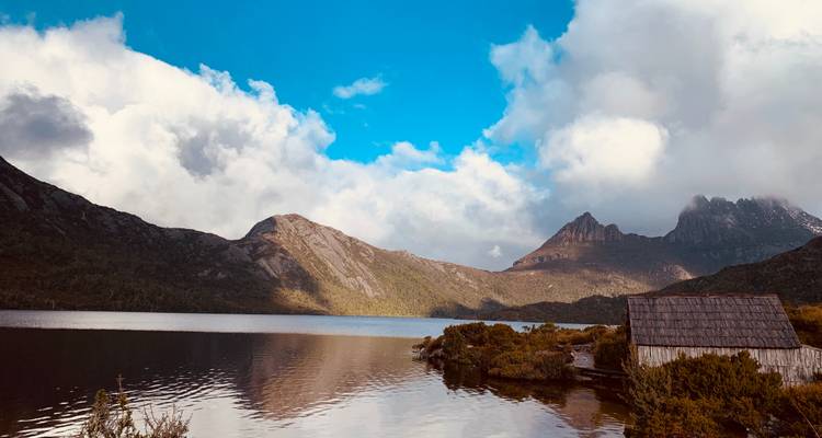 Iconic rugged mountain peaks reflect in a tranquil alpine lake beside an old wooden boatshed